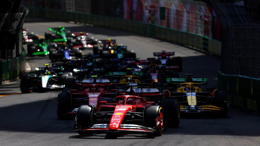 Formula 1 cars, led by a red Ferrari, racing closely together on a narrow street circuit